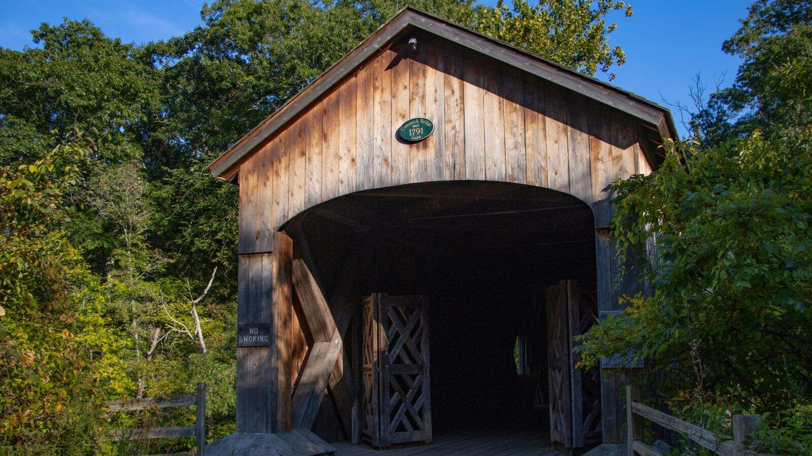 A wooden covered bridge with open doors stands surrounded by green trees under a clear blue sky.