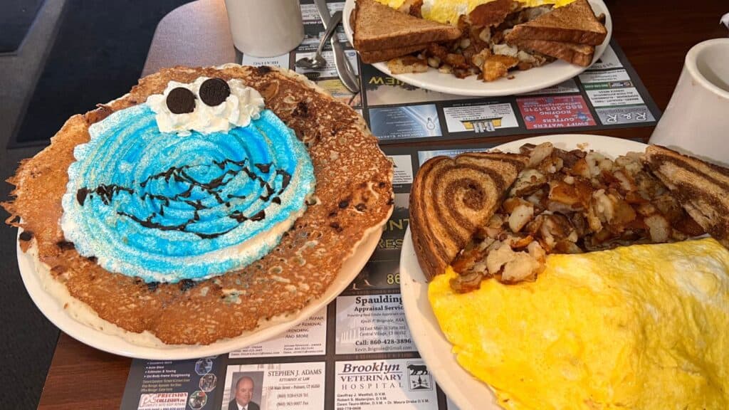 A blue frosted pancake decorated like Cookie Monster with whipped cream and cookies, next to an omelet with toast and home fries on a diner table.