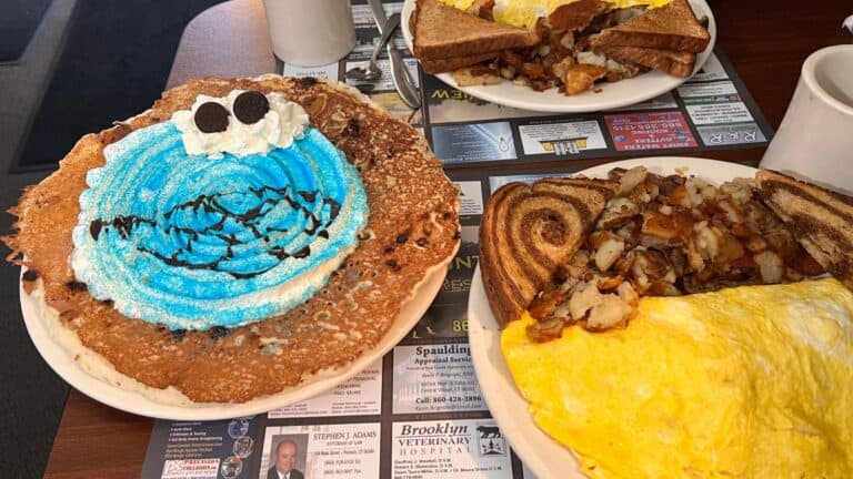 A blue frosted pancake decorated like Cookie Monster with whipped cream and cookies, next to an omelet with toast and home fries on a diner table.