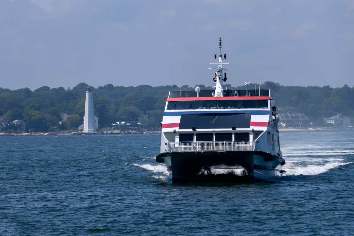 A ferry boat travels across the water toward the camera, with a shoreline, lighthouse, and trees visible in the background under a partly cloudy sky.