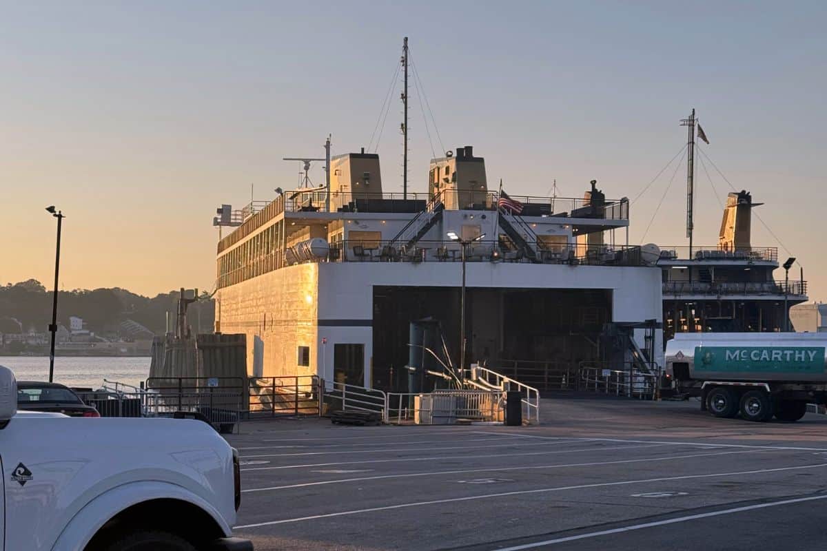 A ferry docked at a terminal during sunrise, with a truck and part of another ship visible nearby.