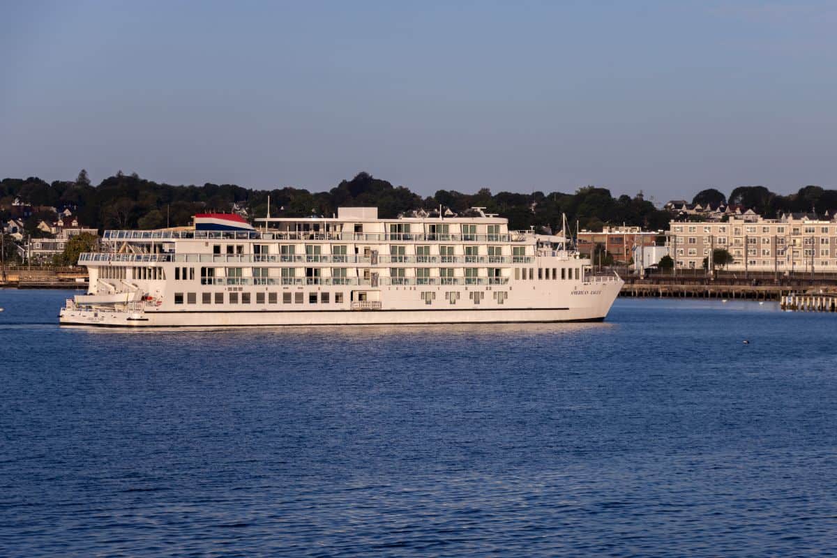 A large white river cruise ship sails near the shoreline with buildings and trees visible in the background under a clear sky.