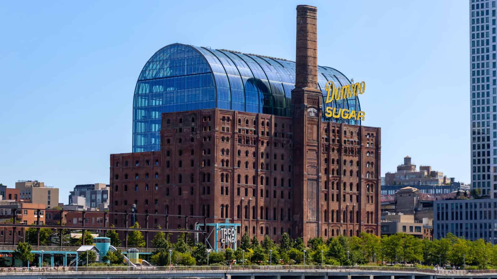 A large brick industrial building with a tall smokestack and a modern glass rooftop addition, featuring a "Domino Sugar" sign, stands among city structures.