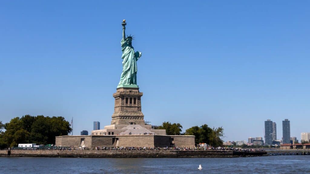The Statue of Liberty stands on Liberty Island in New York Harbor, surrounded by water with city buildings visible in the background.