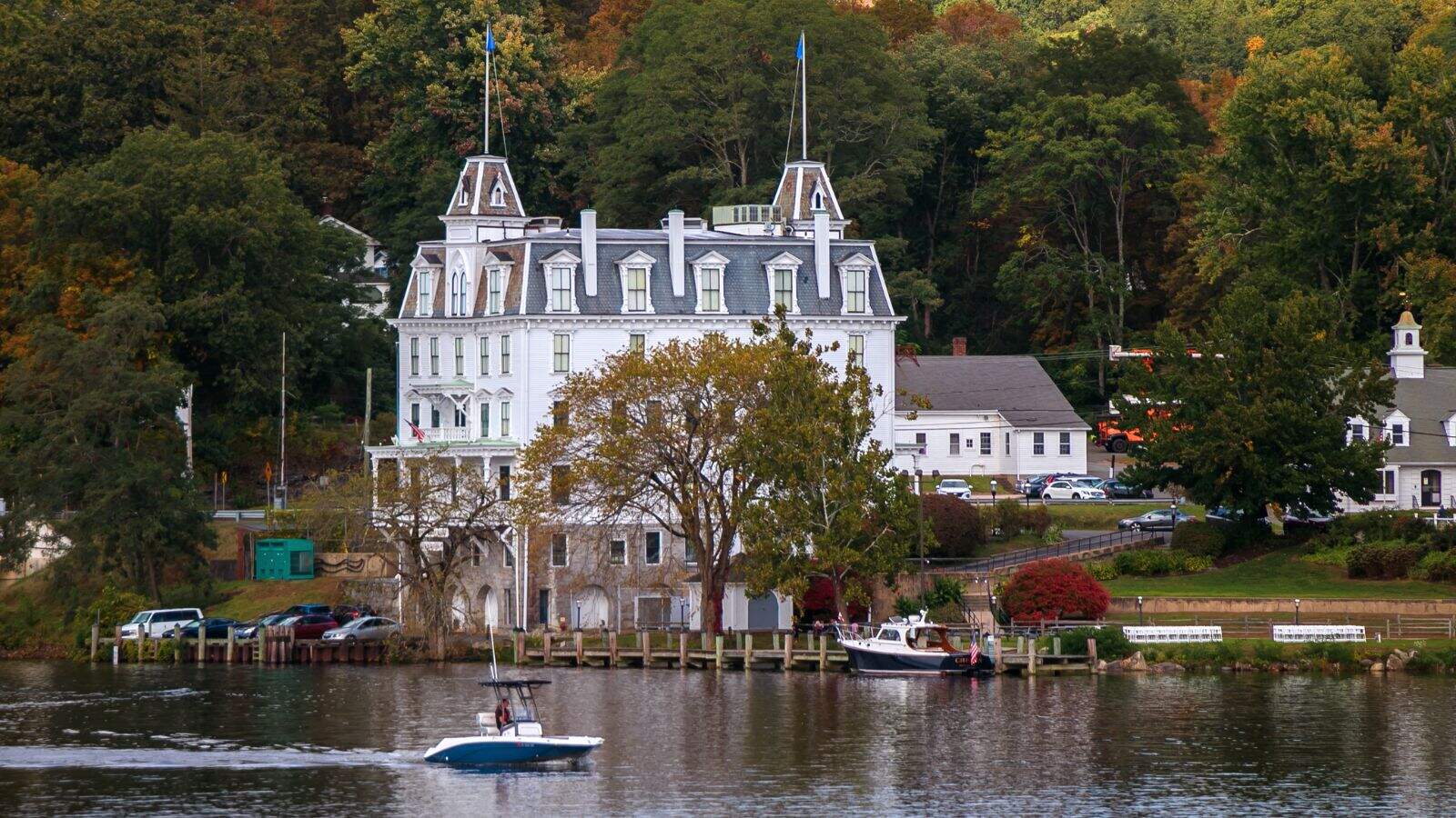 A large white historic building with mansard roof sits by a river, surrounded by trees and smaller buildings; a boat moves on the water in the foreground.