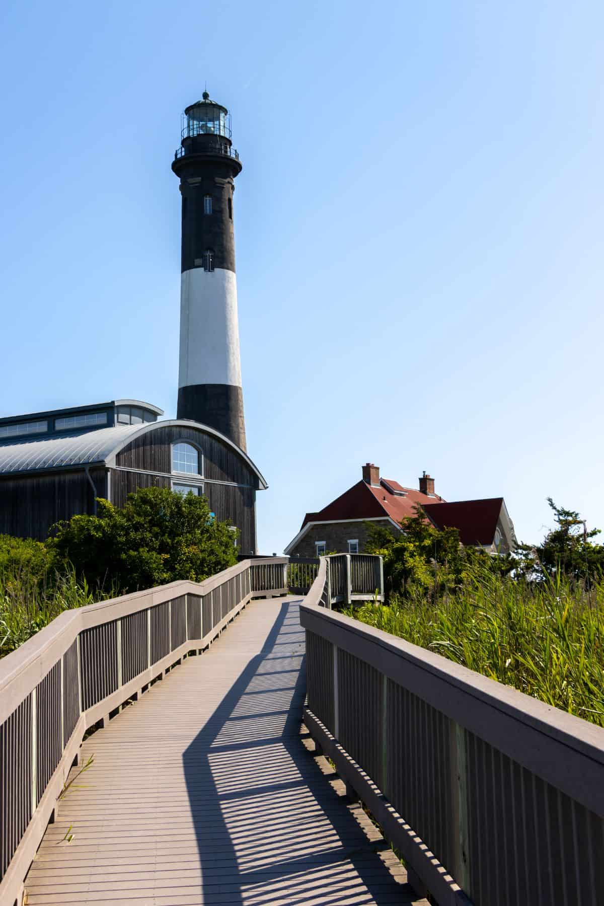 A paved boardwalk leads to a tall black-and-white lighthouse, with a house and greenery nearby under a clear blue sky.