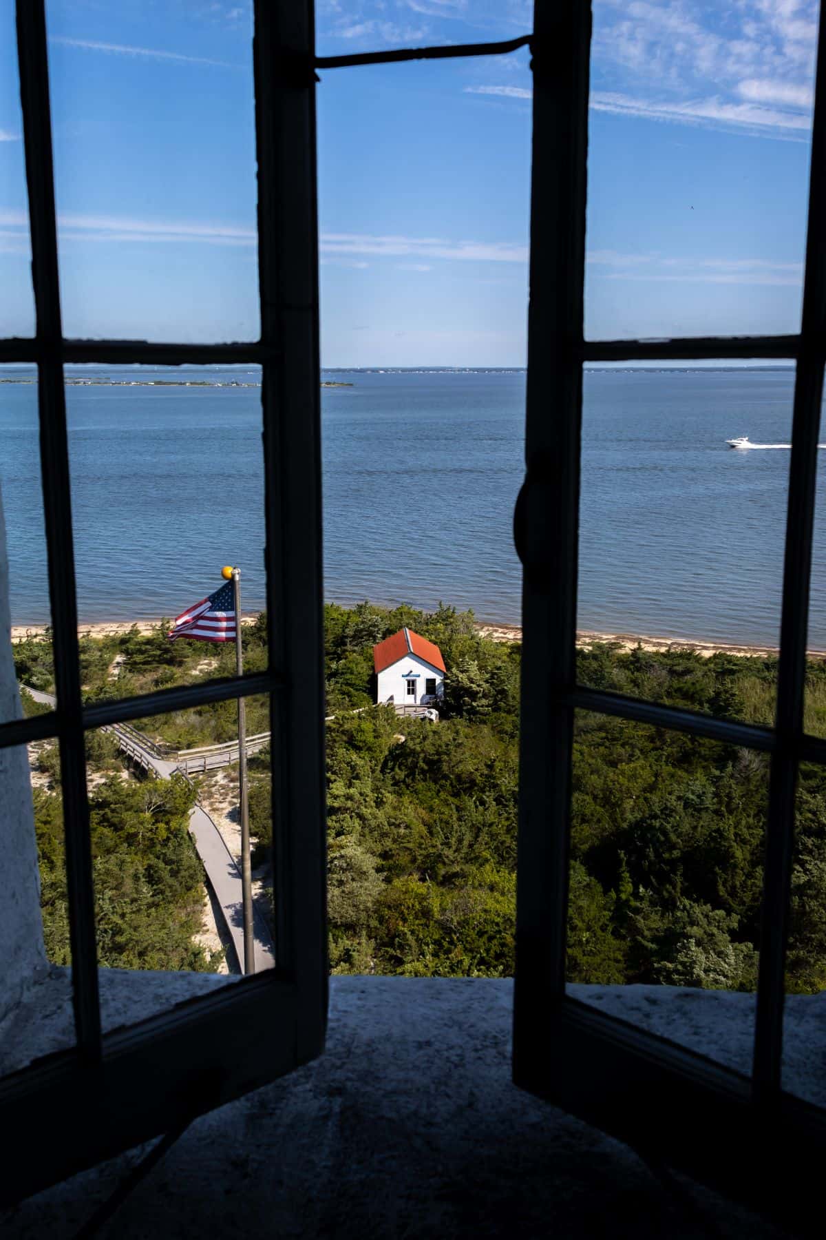 View through an open window showing a small white house with a red roof near the shoreline, a flagpole, greenery, and a body of water under a blue sky.