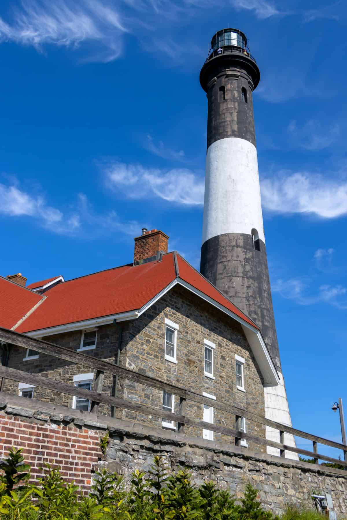 A tall black and white lighthouse stands behind a stone building with a red roof under a blue sky with wispy clouds.