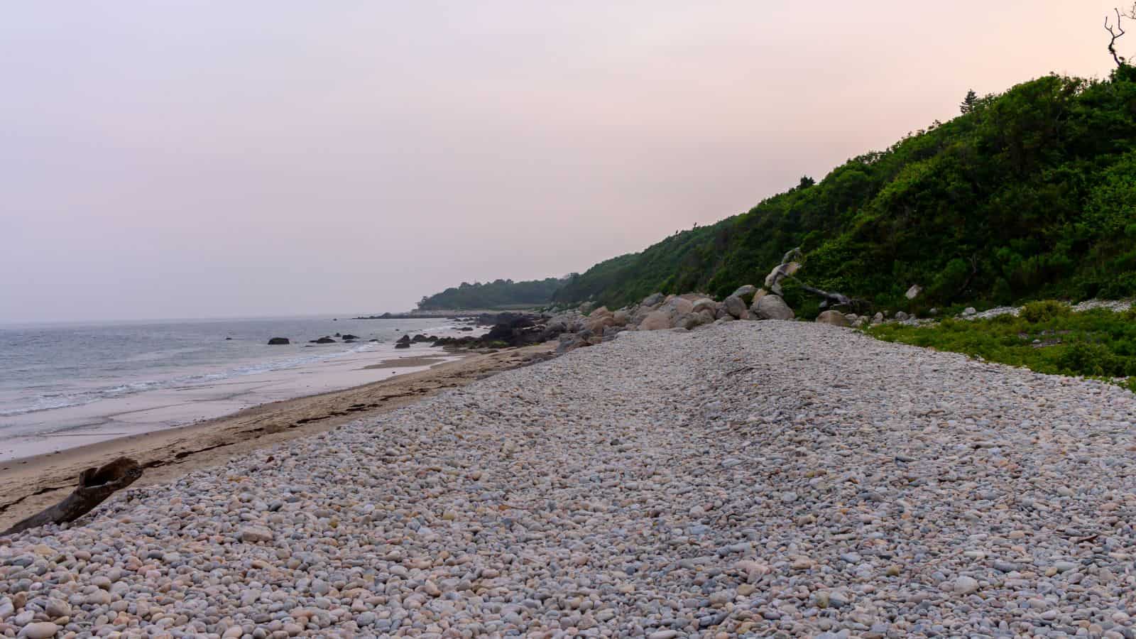 A rocky beach with smooth pebbles and boulders, bordered by green hills, under a cloudy sky near the ocean.