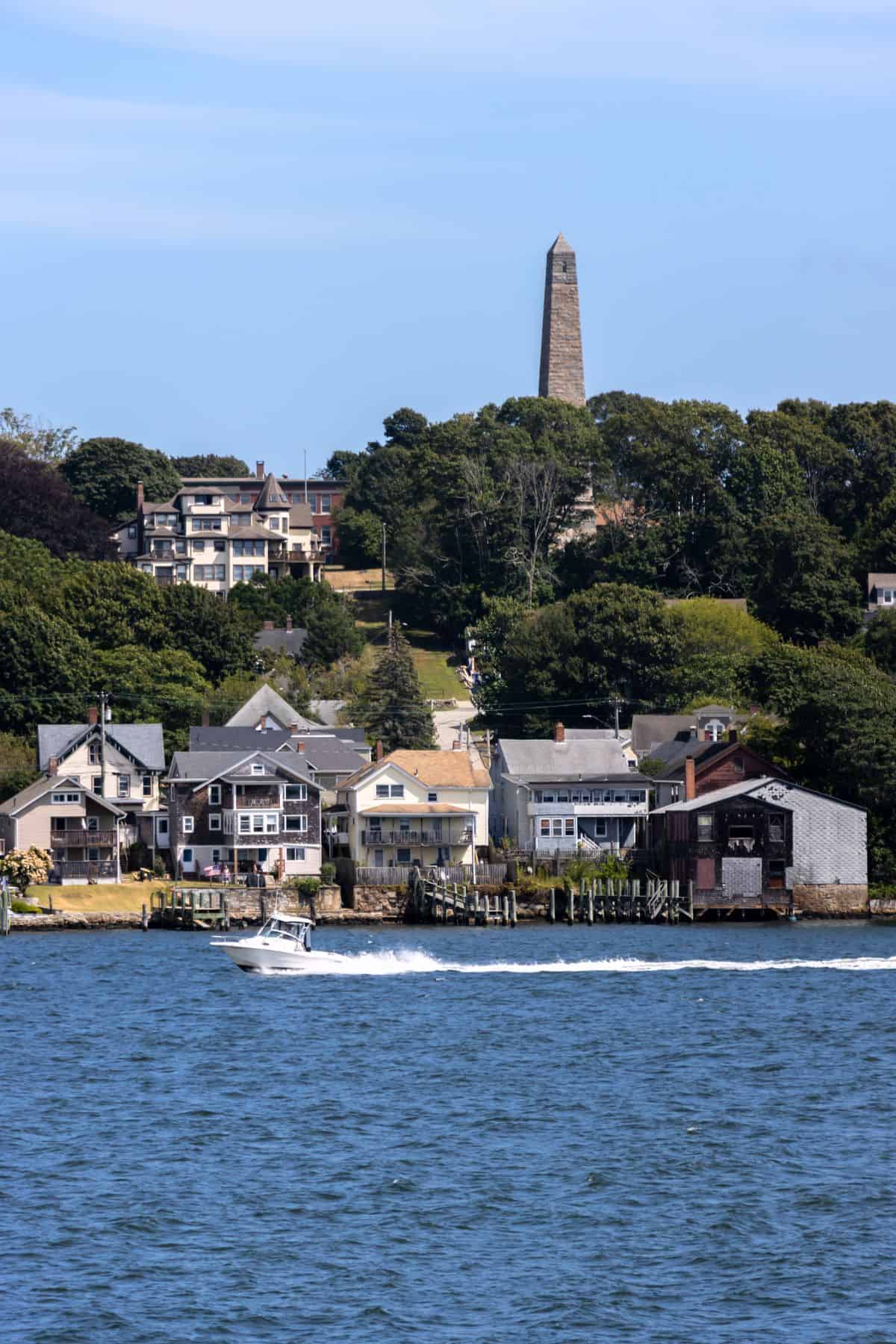 A small motorboat travels on the water in front of waterfront houses, with a tall stone monument on a hill and trees in the background.