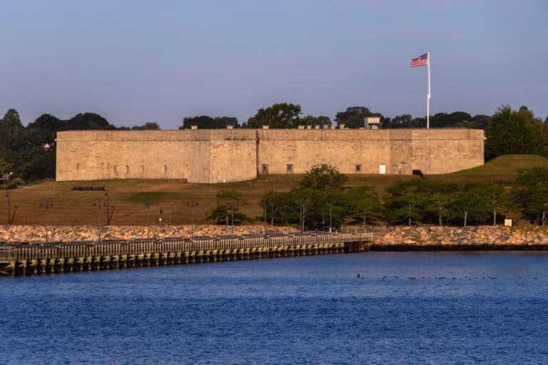 A stone fort with an American flag stands on a grassy hill by the waterfront, with trees and a clear sky in the background.