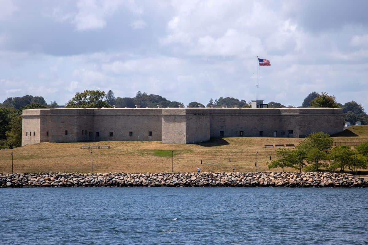 A stone fort sits on grassy land by the water, with an American flag flying above it under a partly cloudy sky.
