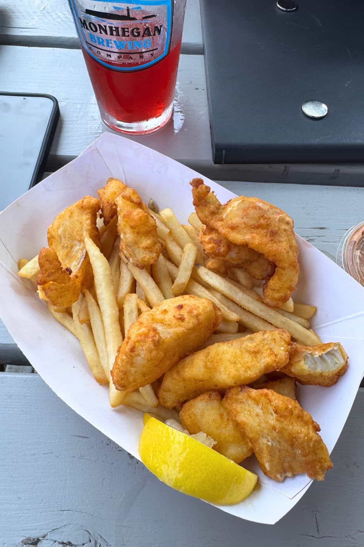 A paper tray filled with fried fish, French fries, and a lemon wedge sits on a picnic table on Monhegan Island, next to a drink and a phone.