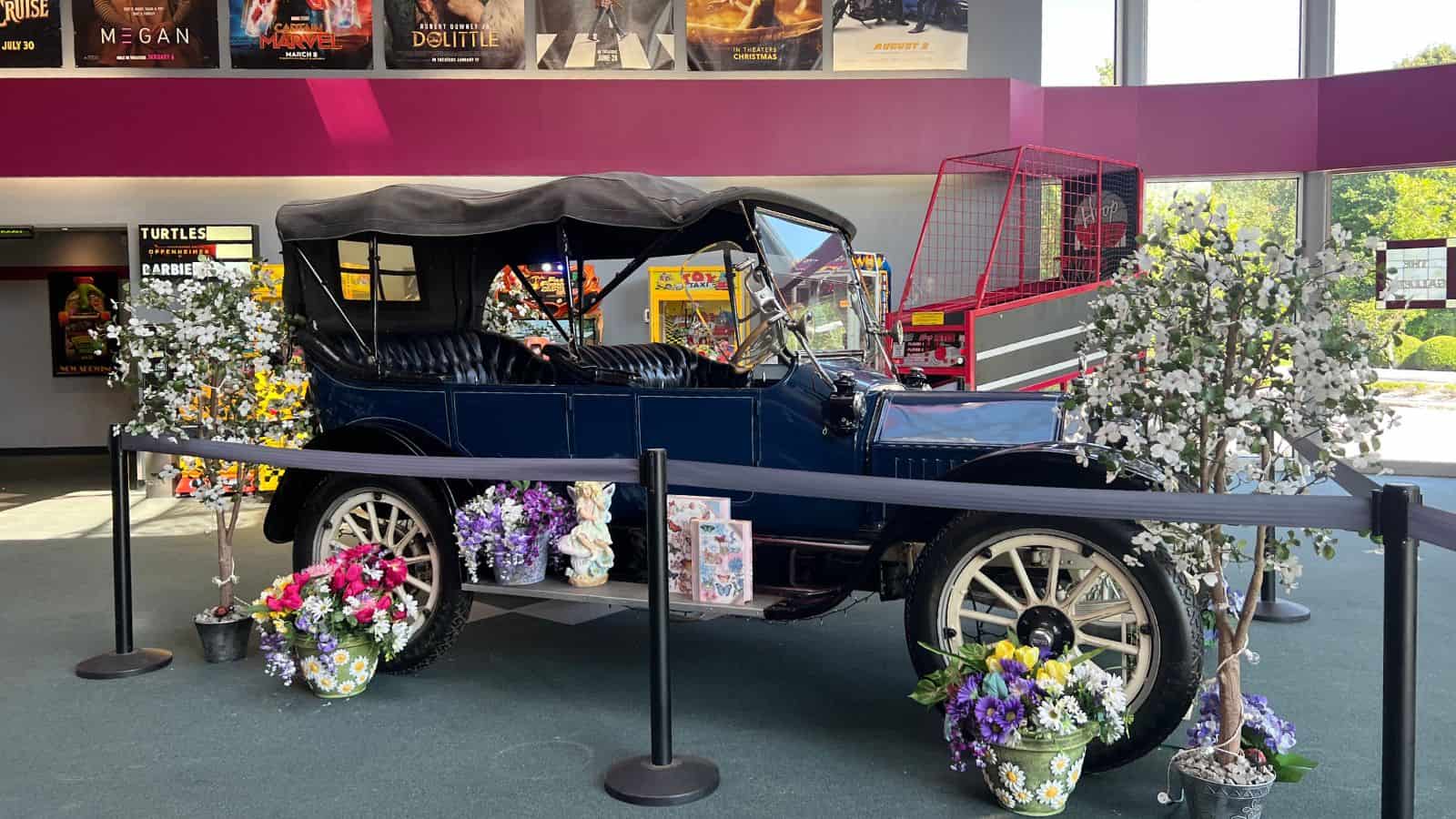 A vintage black car with a soft top is displayed indoors, surrounded by flower arrangements and artificial trees, with movie posters and arcade games in the background.