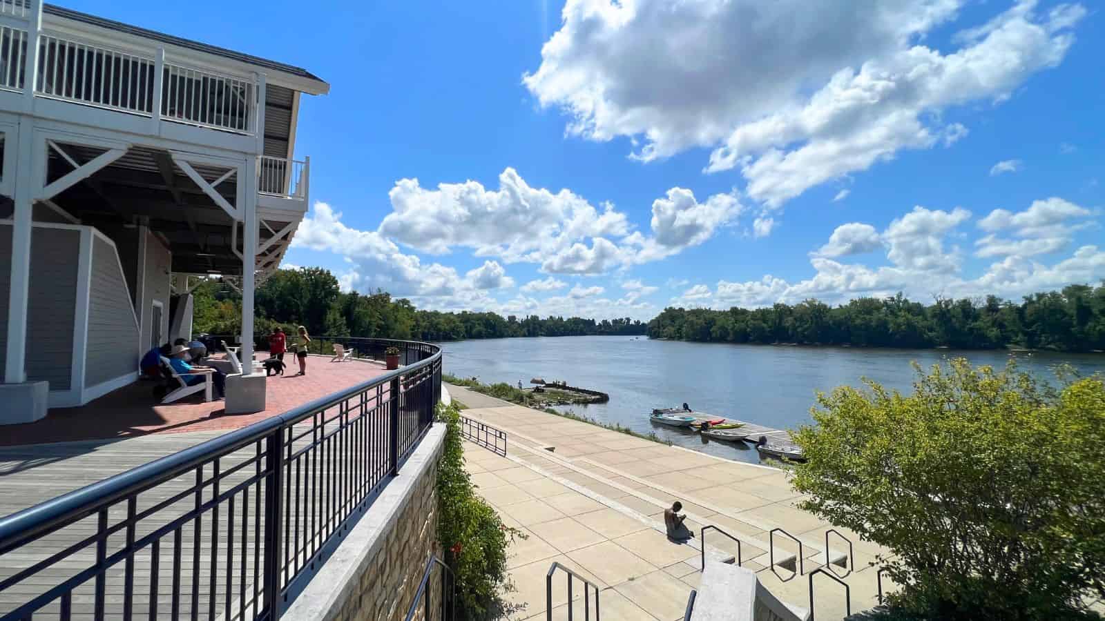 A riverside scene with people sitting near a building, boats docked at the shore, and a partly cloudy sky over the water.