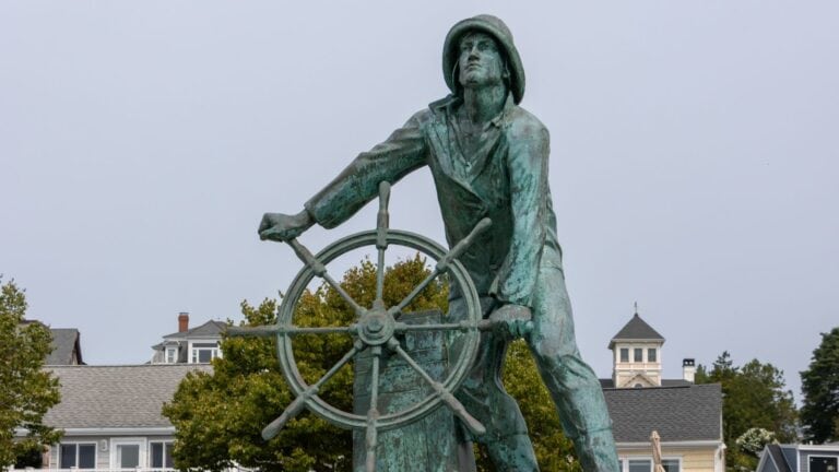 Bronze statue of a fisherman at a ship’s wheel, with buildings and trees in the background under a cloudy sky.