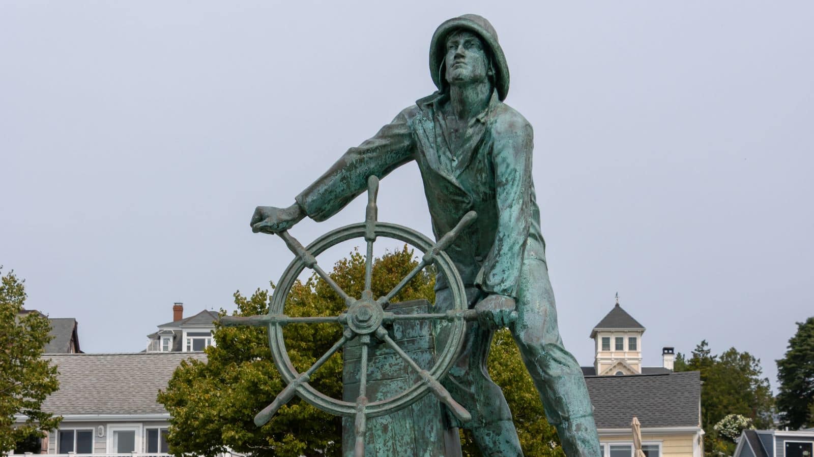 Bronze statue of a fisherman at a ship’s wheel, with buildings and trees in the background under a cloudy sky.
