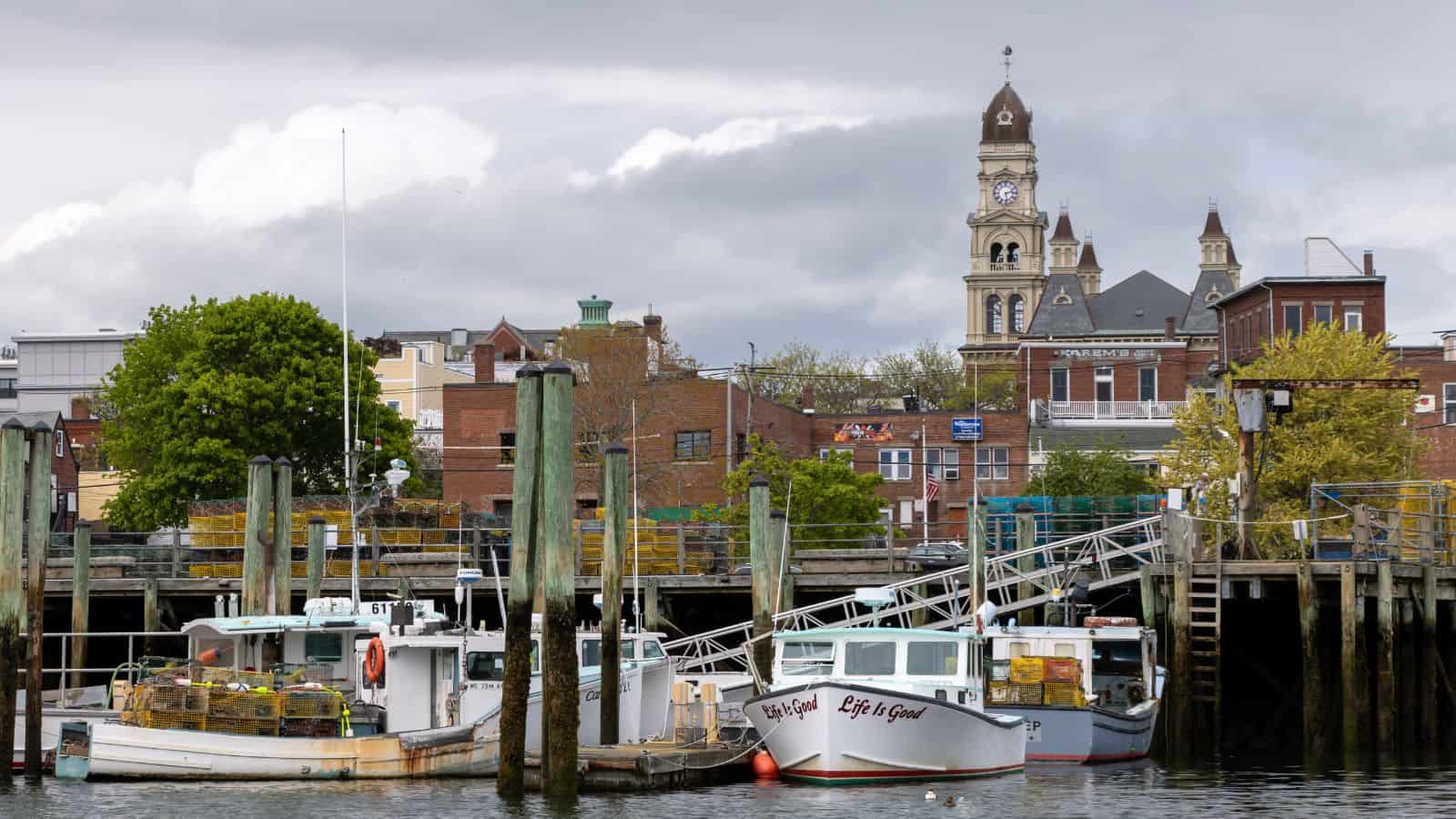 Fishing boats docked at a harbor, with lobster traps on the pier and a clock tower and brick buildings in the background under a cloudy sky.