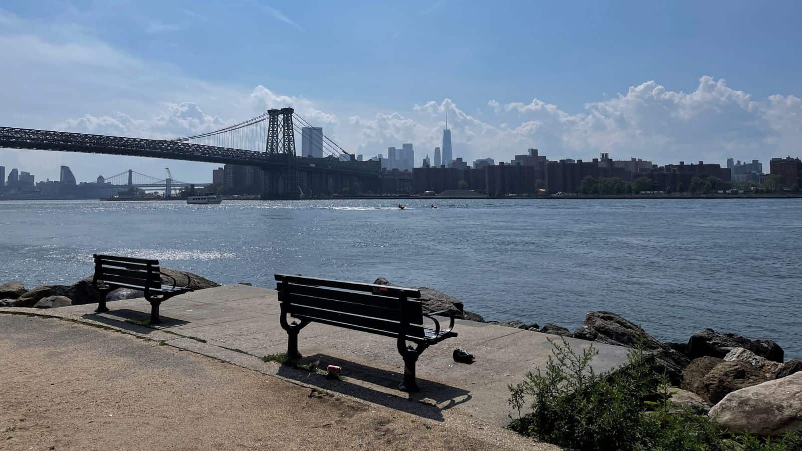Two empty benches face a river with the Williamsburg Bridge and New York City skyline in the background under a partly cloudy sky.