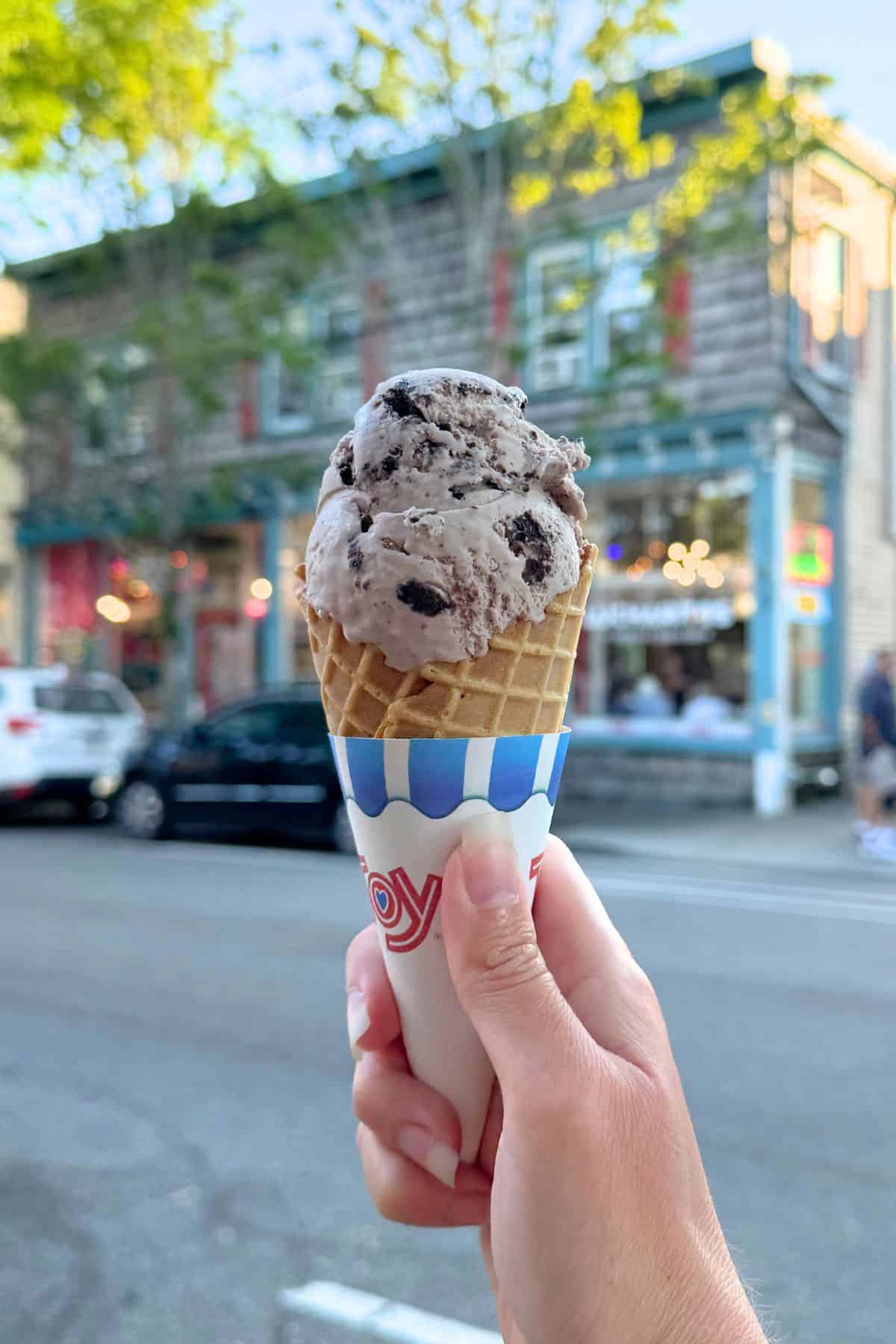 A hand holds an ice cream cone with two scoops of cookies and cream flavor on a street, with a building and parked cars in the background.