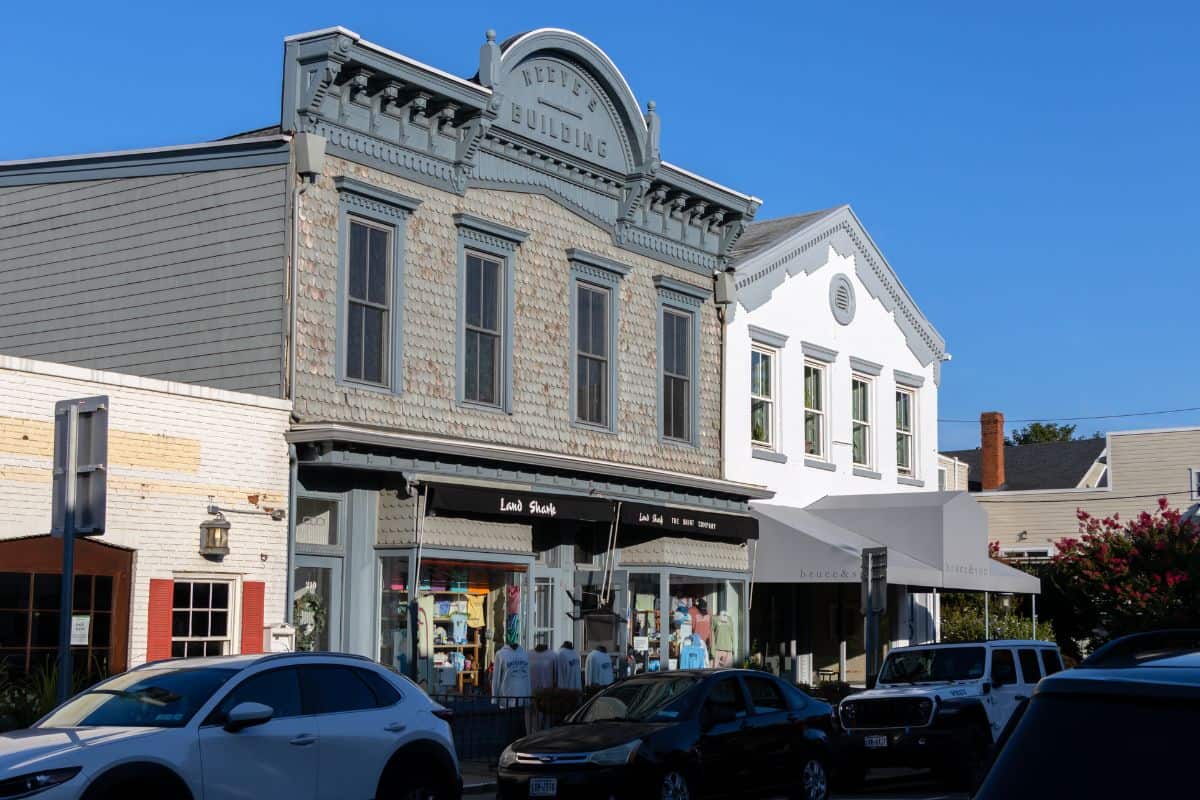 Historic two-story commercial buildings with retail shops on the ground floor, parked cars in front, and a clear blue sky overhead.