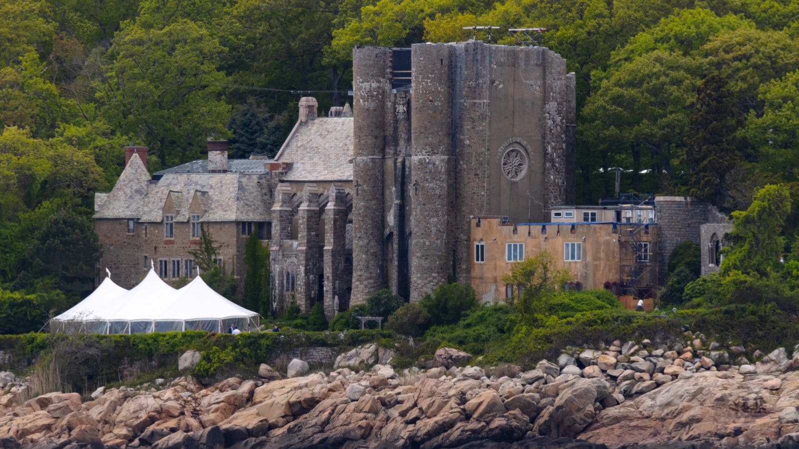 A large stone building with castle-like features sits among trees near a rocky shoreline; a white event tent is set up on the lawn.