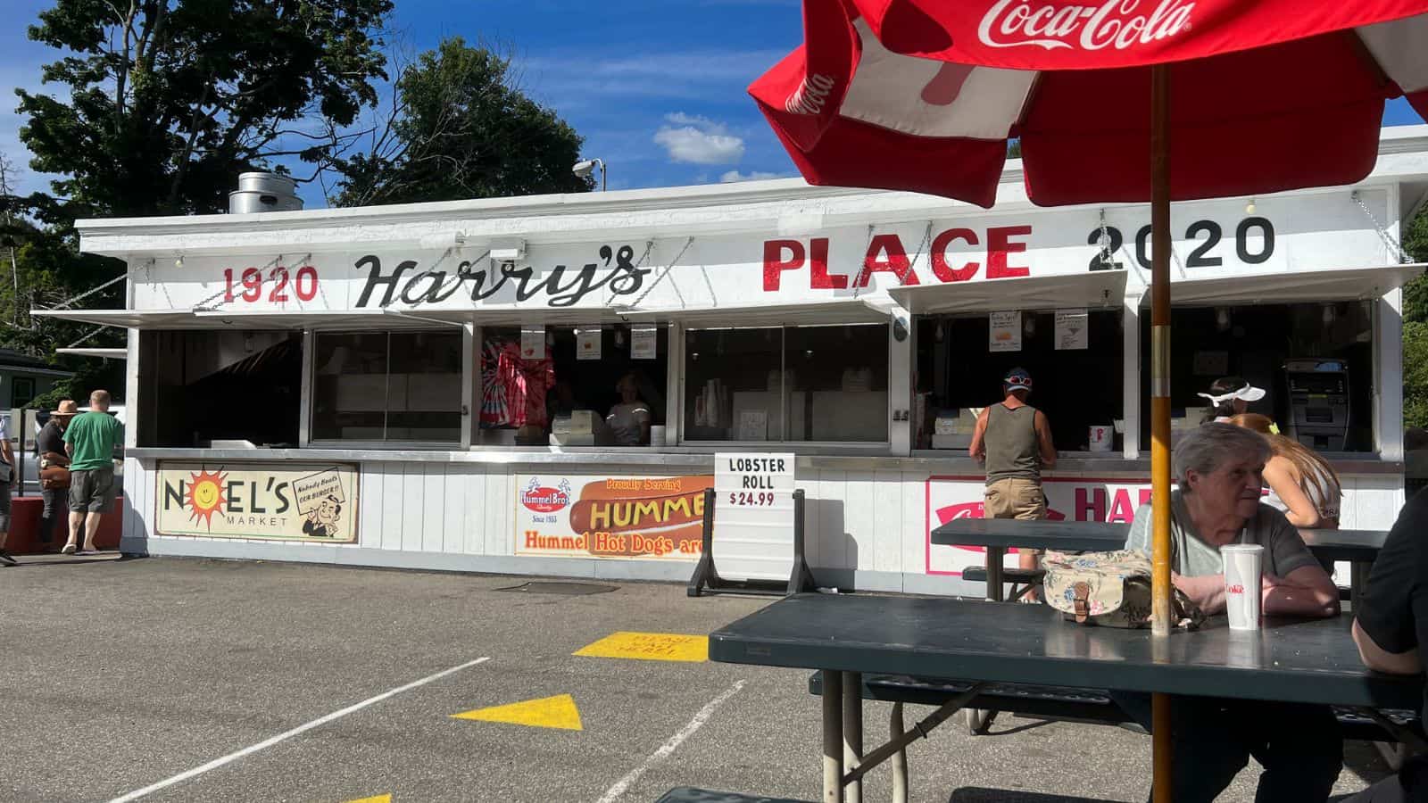 A white roadside food stand labeled "Harry's Place" with a 1920-2020 sign, menu boards, patrons ordering, and people sitting at picnic tables under a Coca-Cola umbrella.