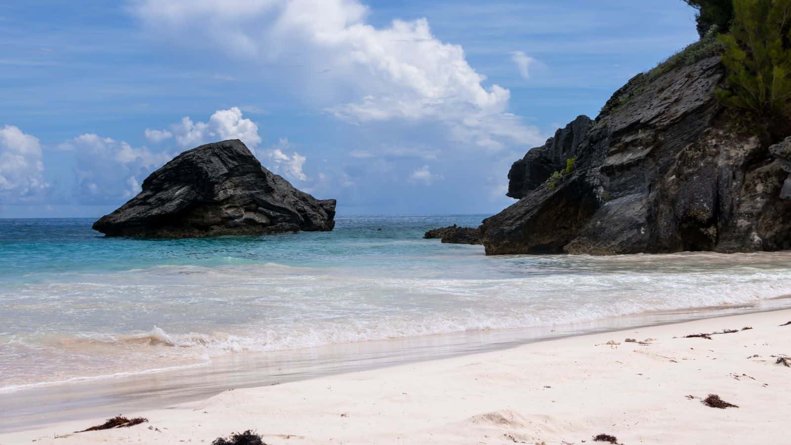 View of a sandy beach with seaweed, turquoise water, rocky cliffs, and a large rock formation in the shallow ocean under a partly cloudy sky.