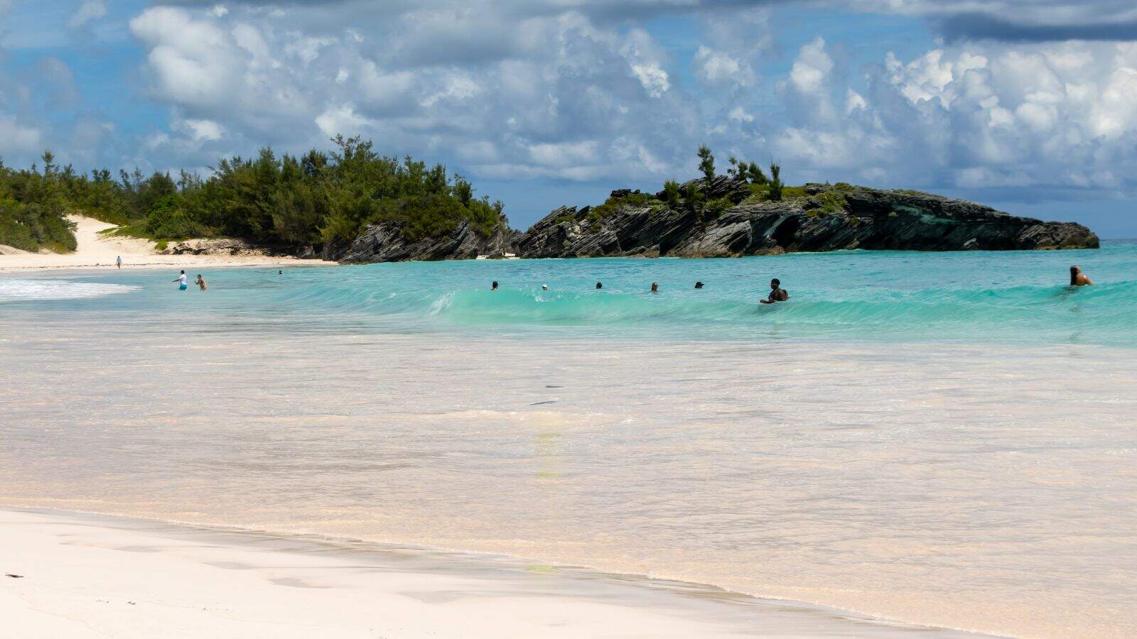 People swimming in clear turquoise water near a sandy beach with a rocky, tree-covered island in the background under a partly cloudy sky.
