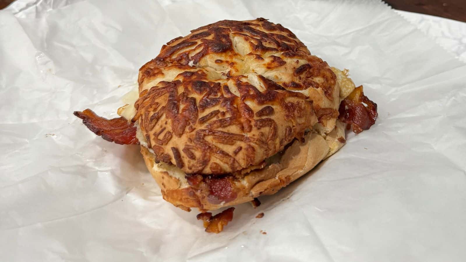 A sandwich with a cheese-crusted bun, bacon, and other fillings sits on a sheet of crumpled white parchment paper.