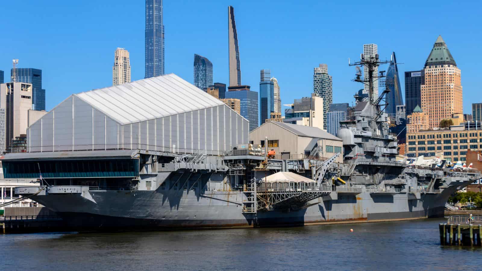 A large gray aircraft carrier is docked at a city waterfront, with modern high-rise buildings in the background under a clear blue sky.