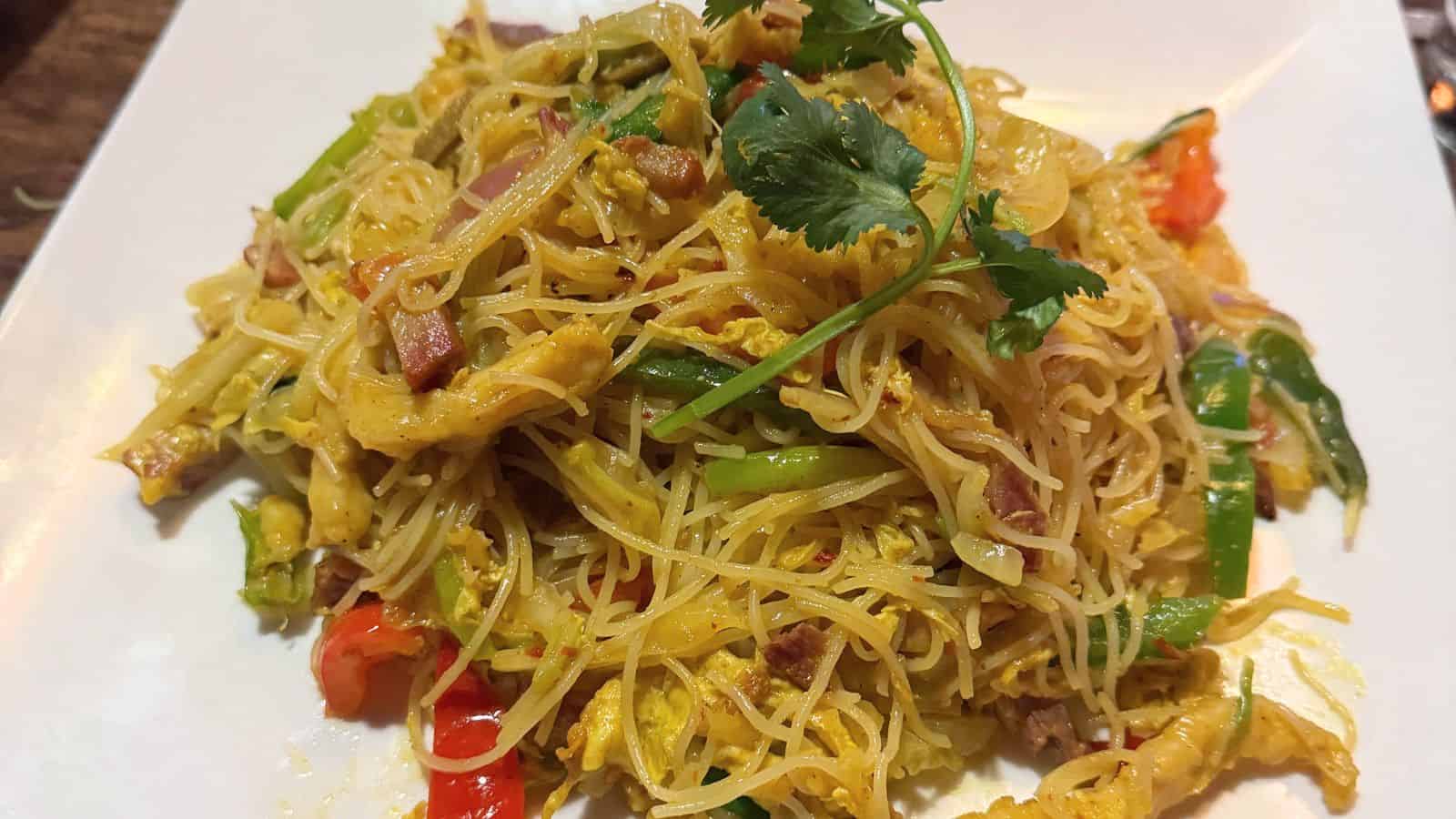 A plate of stir-fried vermicelli noodles with vegetables, scrambled eggs, and meat, garnished with a sprig of cilantro.