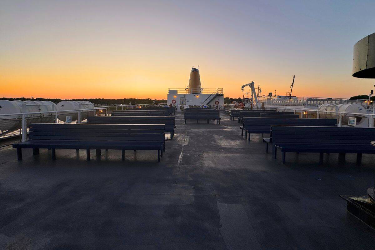 Empty benches on the deck of a ferry at sunset, with life rafts on the sides and trees visible in the distance.