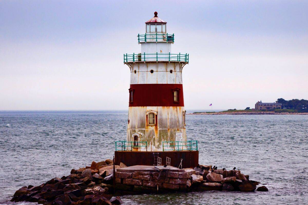 A weathered lighthouse with red and white bands stands on a rocky outcrop surrounded by water under a cloudy sky.