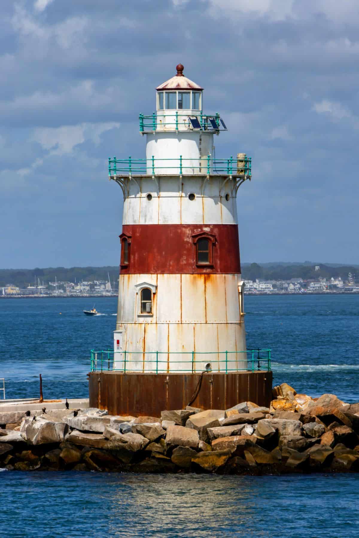A cylindrical lighthouse with a red and white stripes stands on a rocky breakwater surrounded by blue water, with a distant shoreline in the background.
