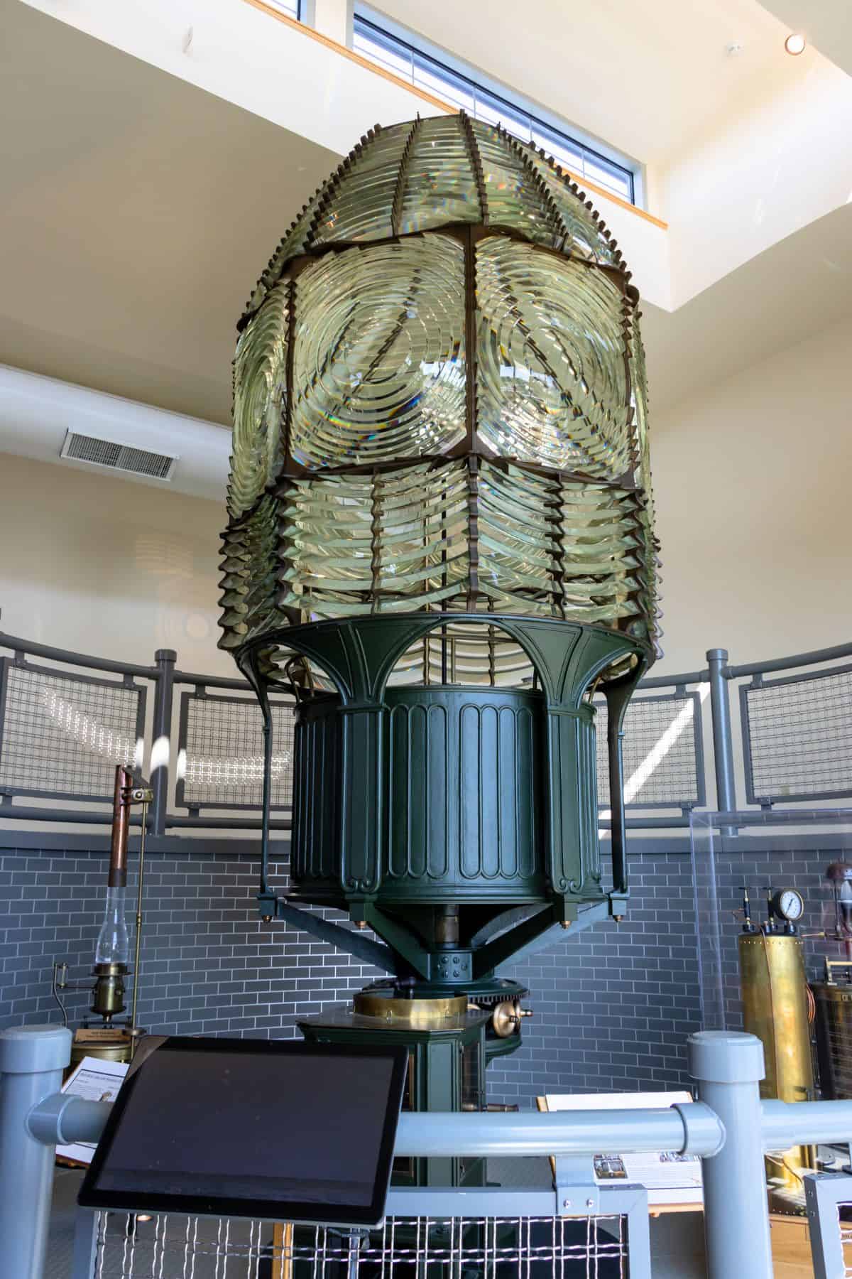 Large antique Fresnel lens from a lighthouse displayed indoors behind a safety barrier, with an informational panel in front.