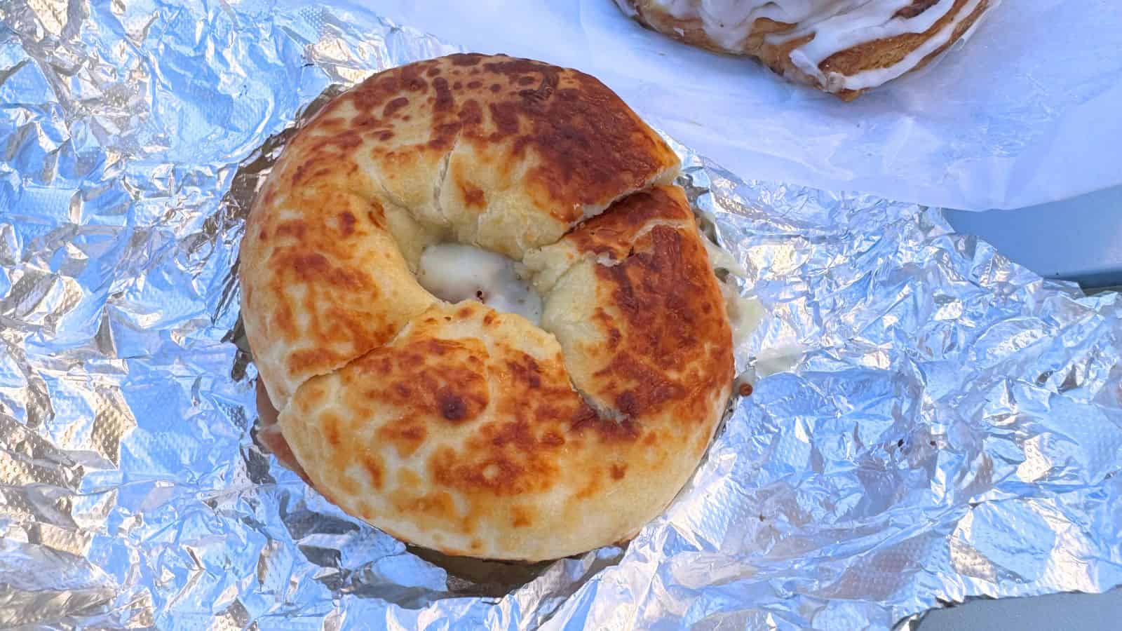 A close-up of a grilled bagel sandwich with melted cheese, placed on crumpled aluminum foil.