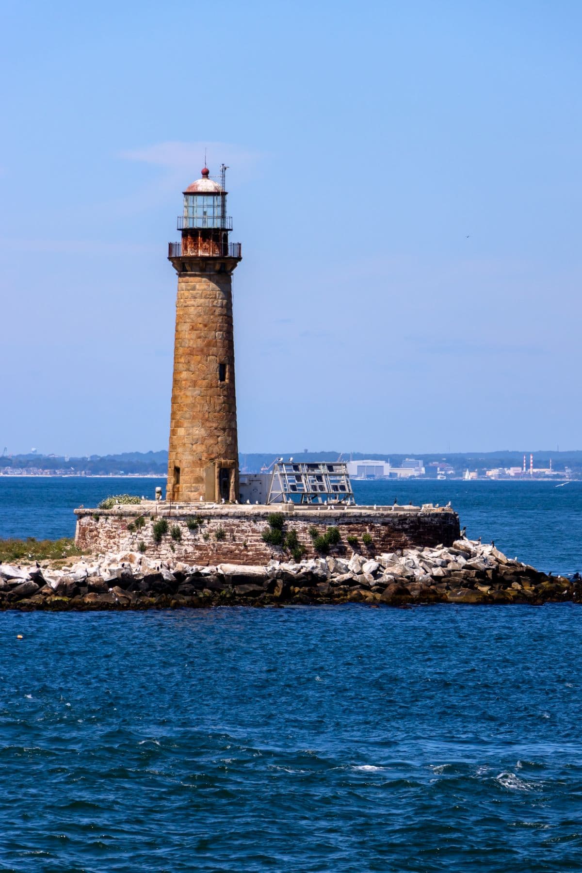 A tall stone lighthouse stands on a rocky island surrounded by blue water, with buildings and distant land visible in the background.