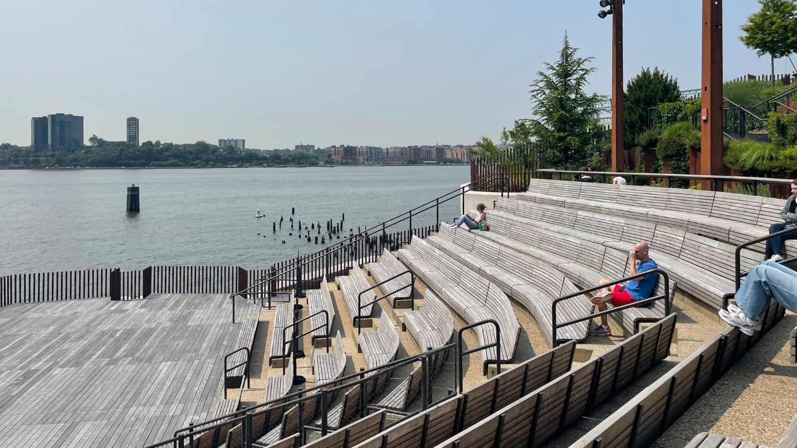 Outdoor wooden amphitheater seating overlooks a river with a few people relaxing, city buildings visible across the water under a clear sky—one of the best things to do along the water in NYC.