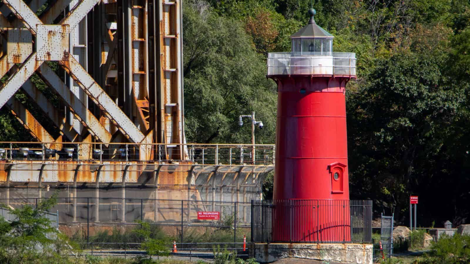 A small red lighthouse stands near a rusted metal bridge structure, surrounded by trees and fenced off from the public.