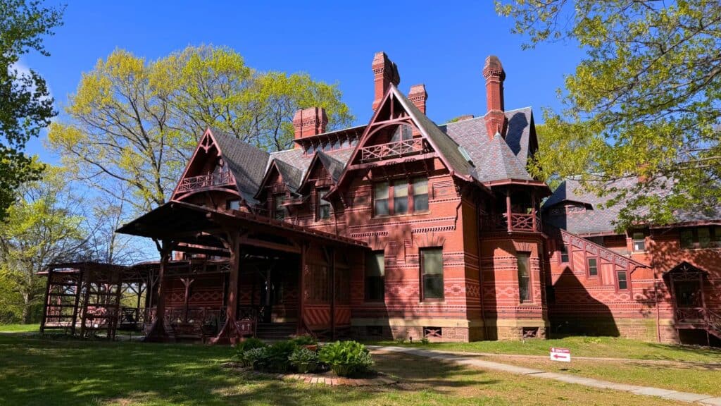 A large red Victorian-style house with intricate woodwork sits surrounded by trees under a clear blue sky.