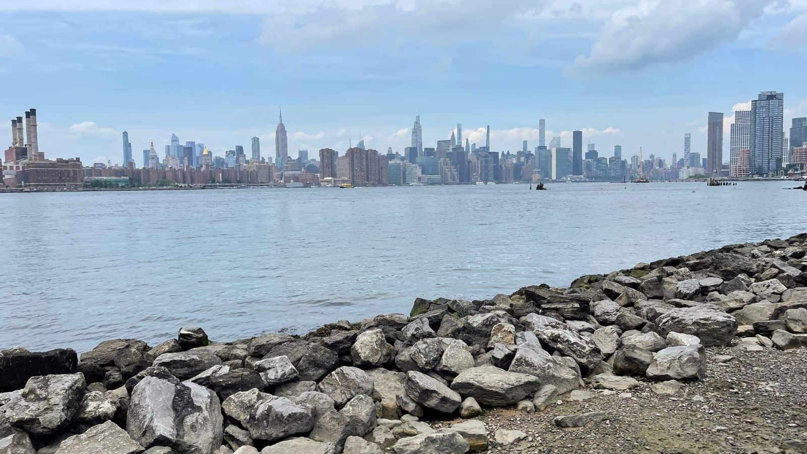 Rocky shoreline in the foreground with the East River and New York City skyline, including the Empire State Building, in the background under a partly cloudy sky.