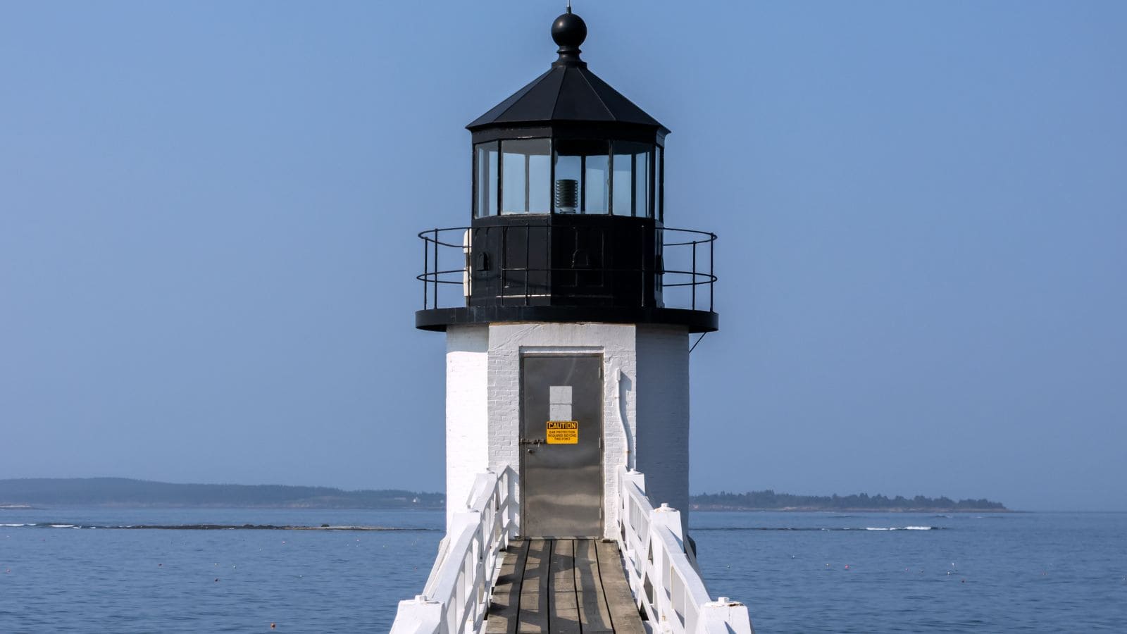 A white lighthouse with a black top stands at the end of a wooden walkway over the water; a yellow warning sign is posted on its door.