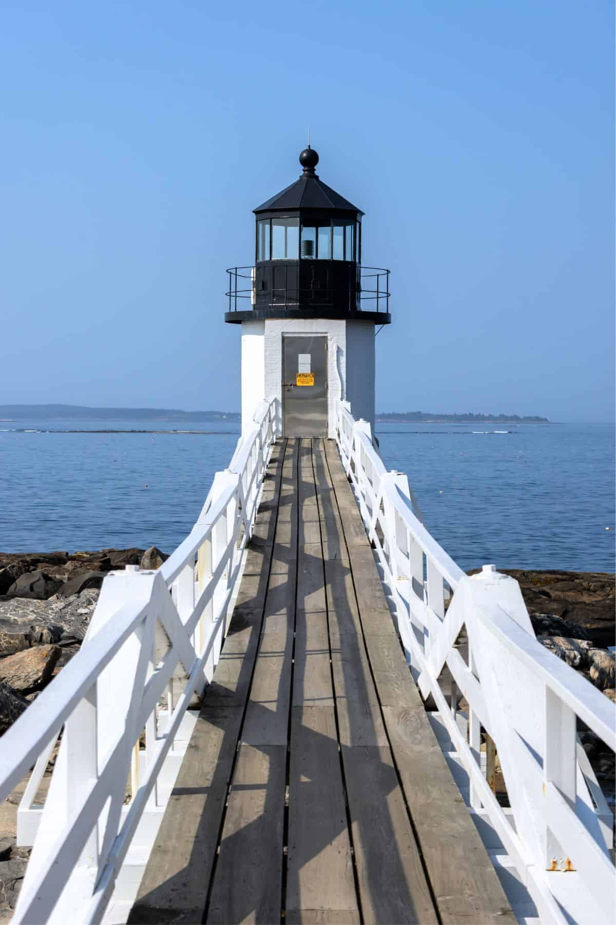 A wooden walkway leads to a small white lighthouse with a black top, situated on a rocky shoreline against a calm blue sea.