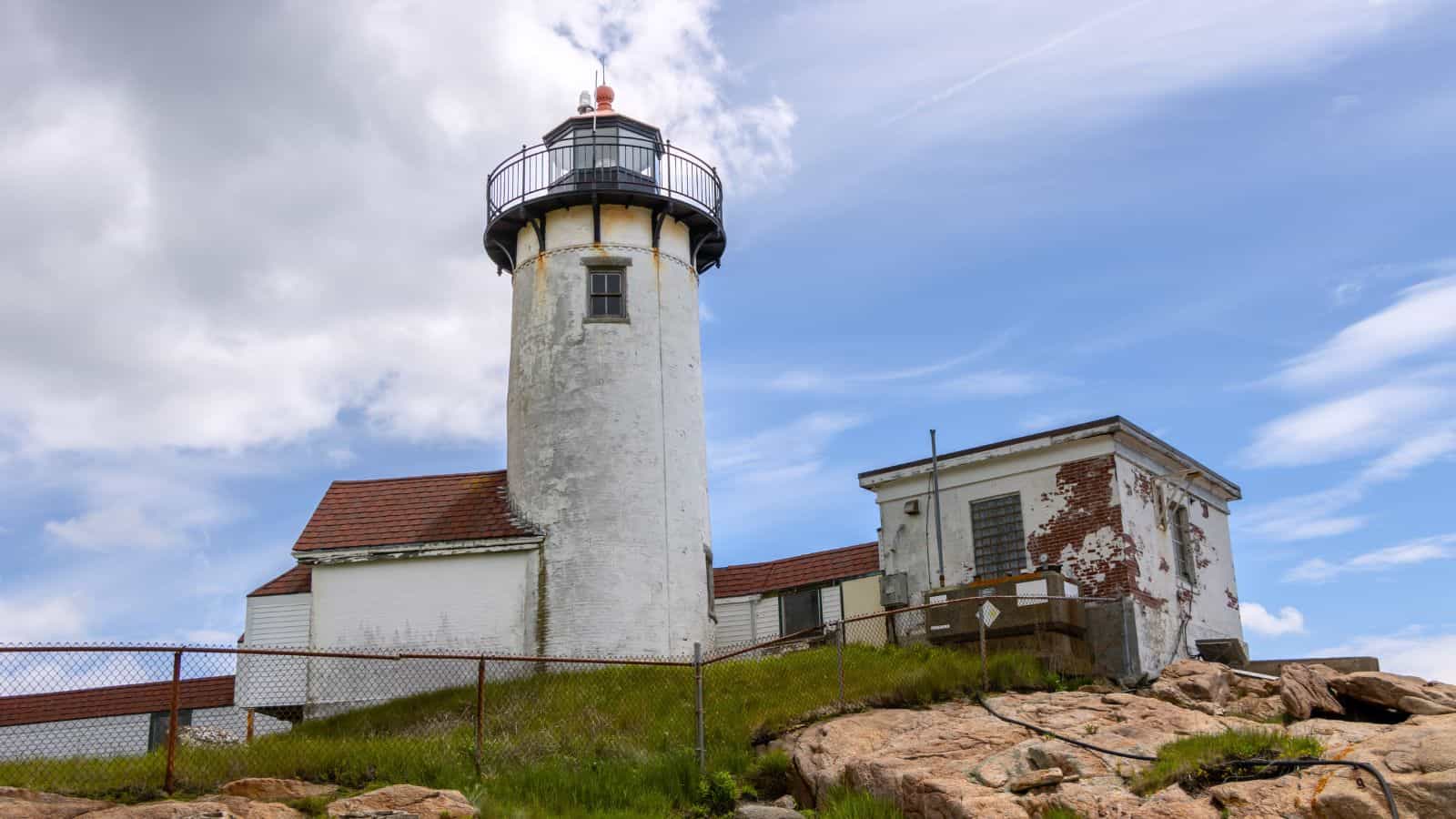 A weathered white lighthouse with an attached building stands on rocky terrain, surrounded by a metal fence under a partly cloudy sky.
