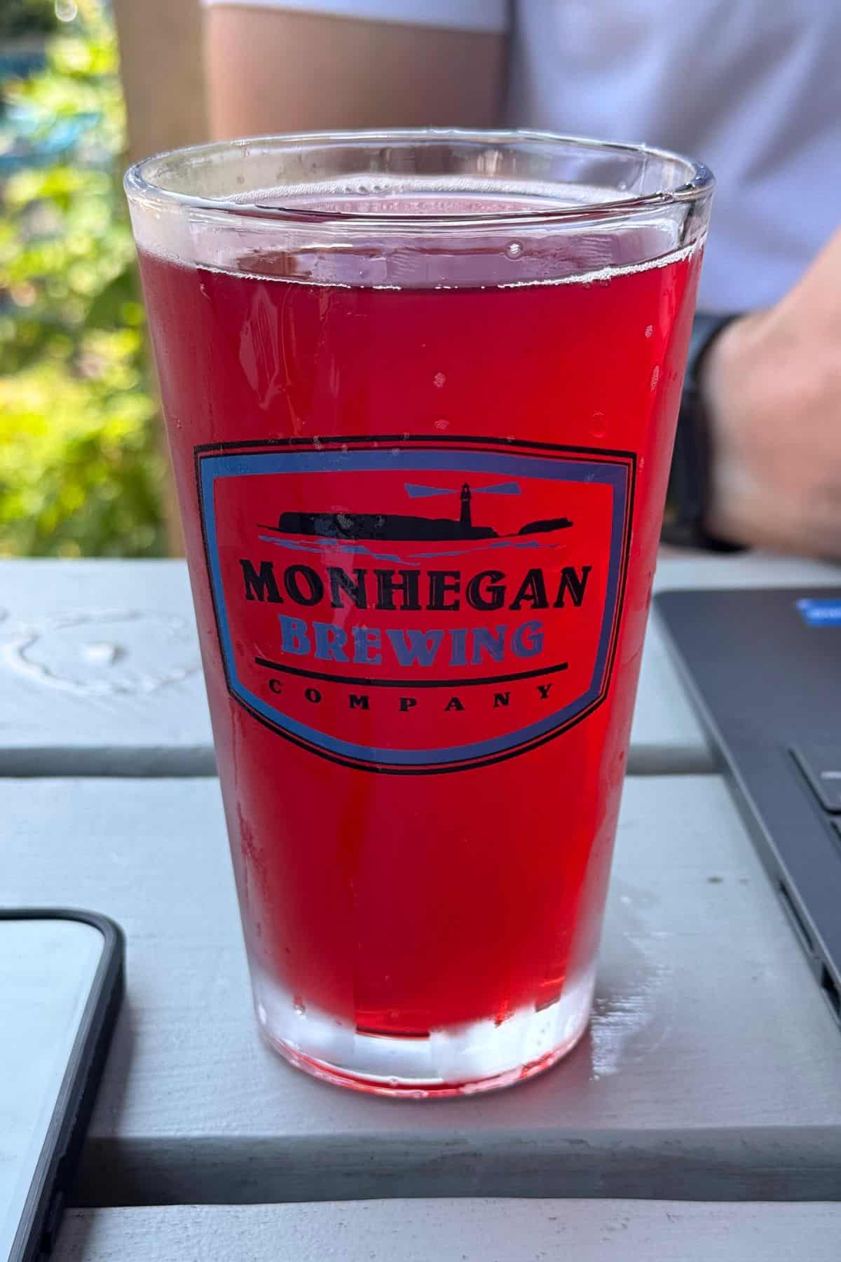 A pint glass filled with a red beverage sits on a wooden table, displaying the Monhegan Brewing Company logo—a nod to Monhegan Island. A phone and laptop are partially visible nearby.