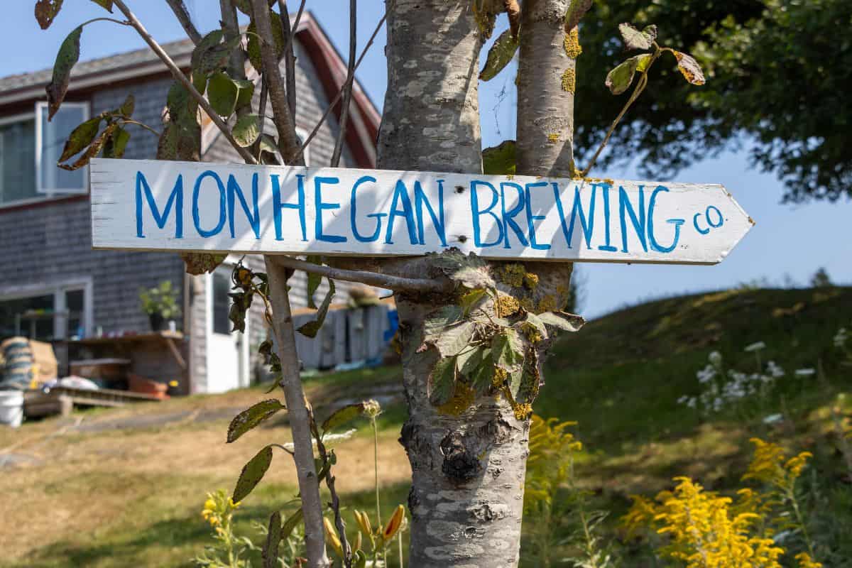A wooden sign reading "Monhegan Brewing Co." is nailed to a tree on Monhegan Island, with a house, greenery, and yellow flowers in the background.