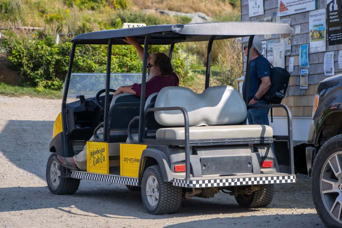A yellow and black golf cart labeled "taxi" is parked outdoors on Monhegan Island; a driver sits inside while a passenger stands nearby next to a bulletin board.
