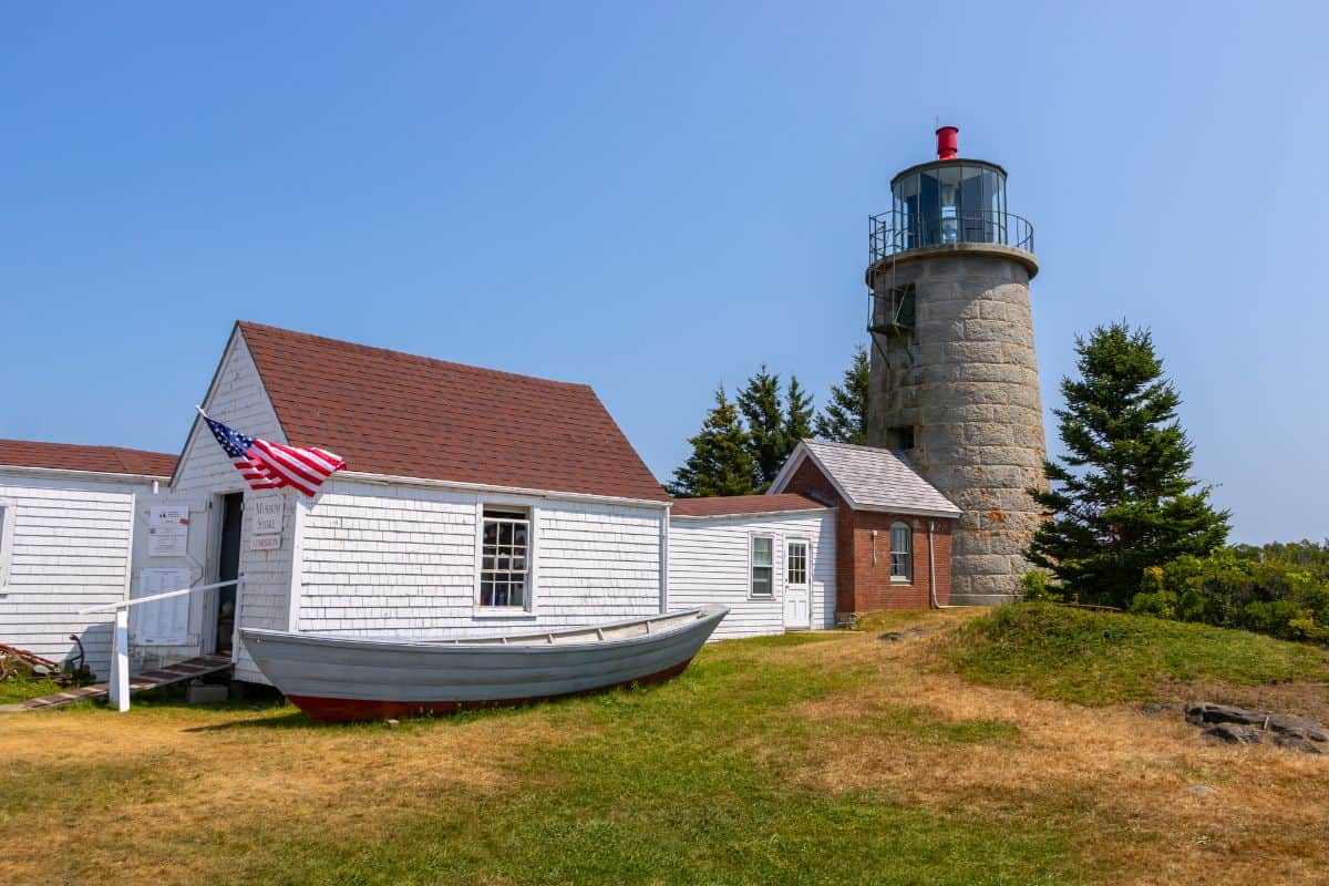 A white wooden building with an American flag stands next to a small boat and a stone lighthouse on Monhegan Island, set atop a grassy hill under a clear blue sky.