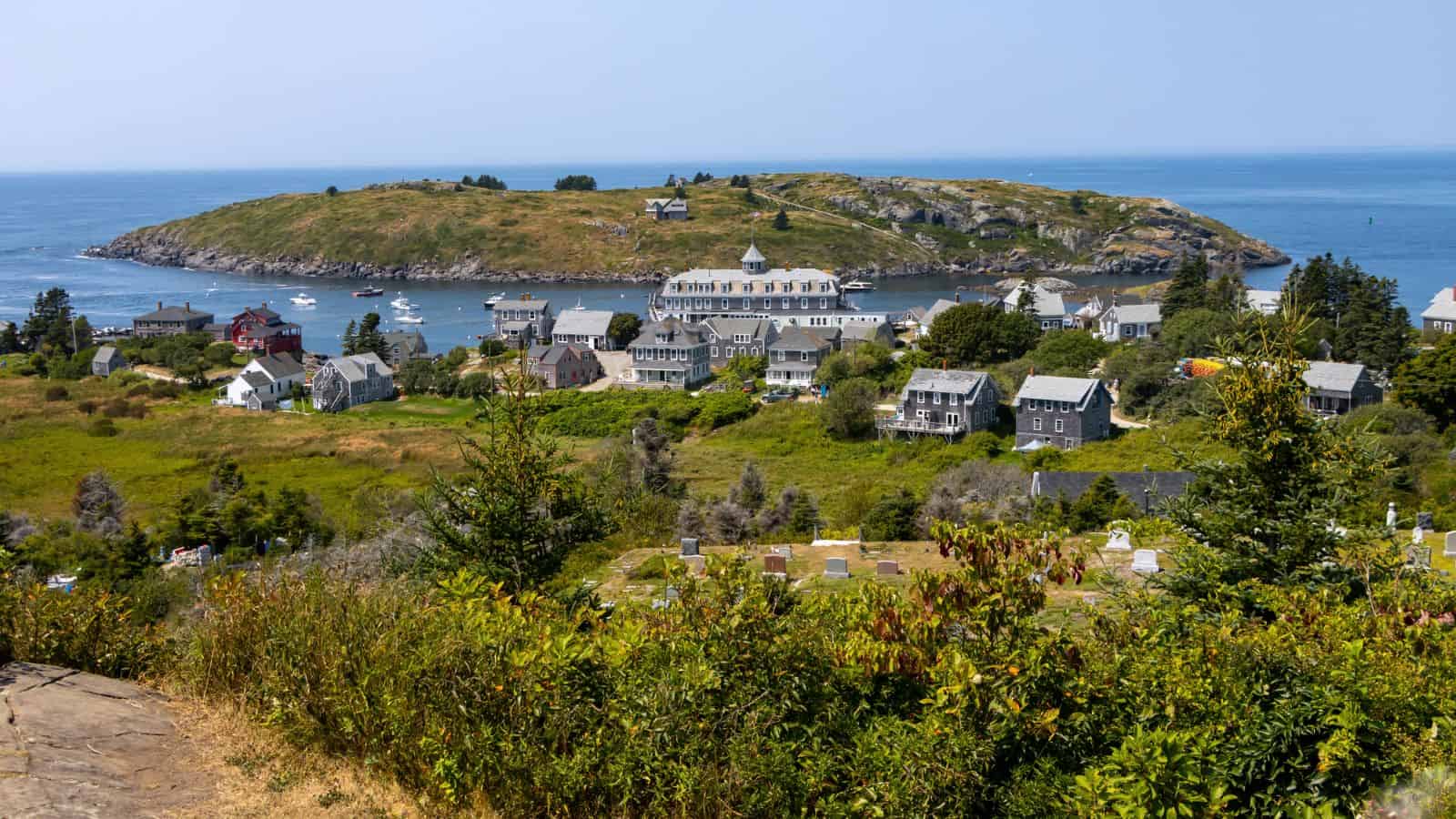 A coastal village with grey houses sits by the water, with boats docked near shore and a green, hilly island in the background under a clear sky.